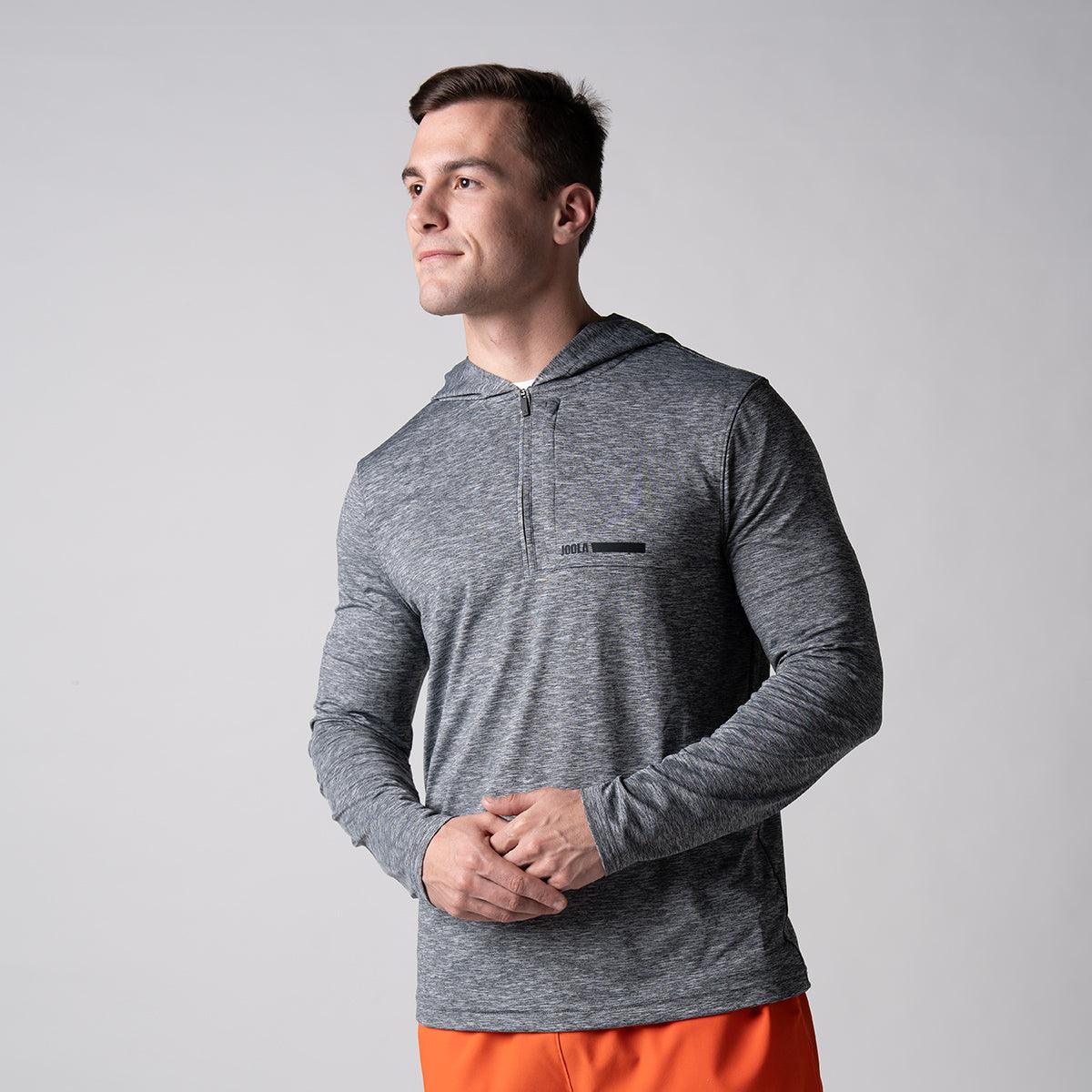 Mann trägt graues, langärmeliges Funktionsshirt mit halbem Reißverschluss und Kapuze, kombiniert mit orangen Shorts.