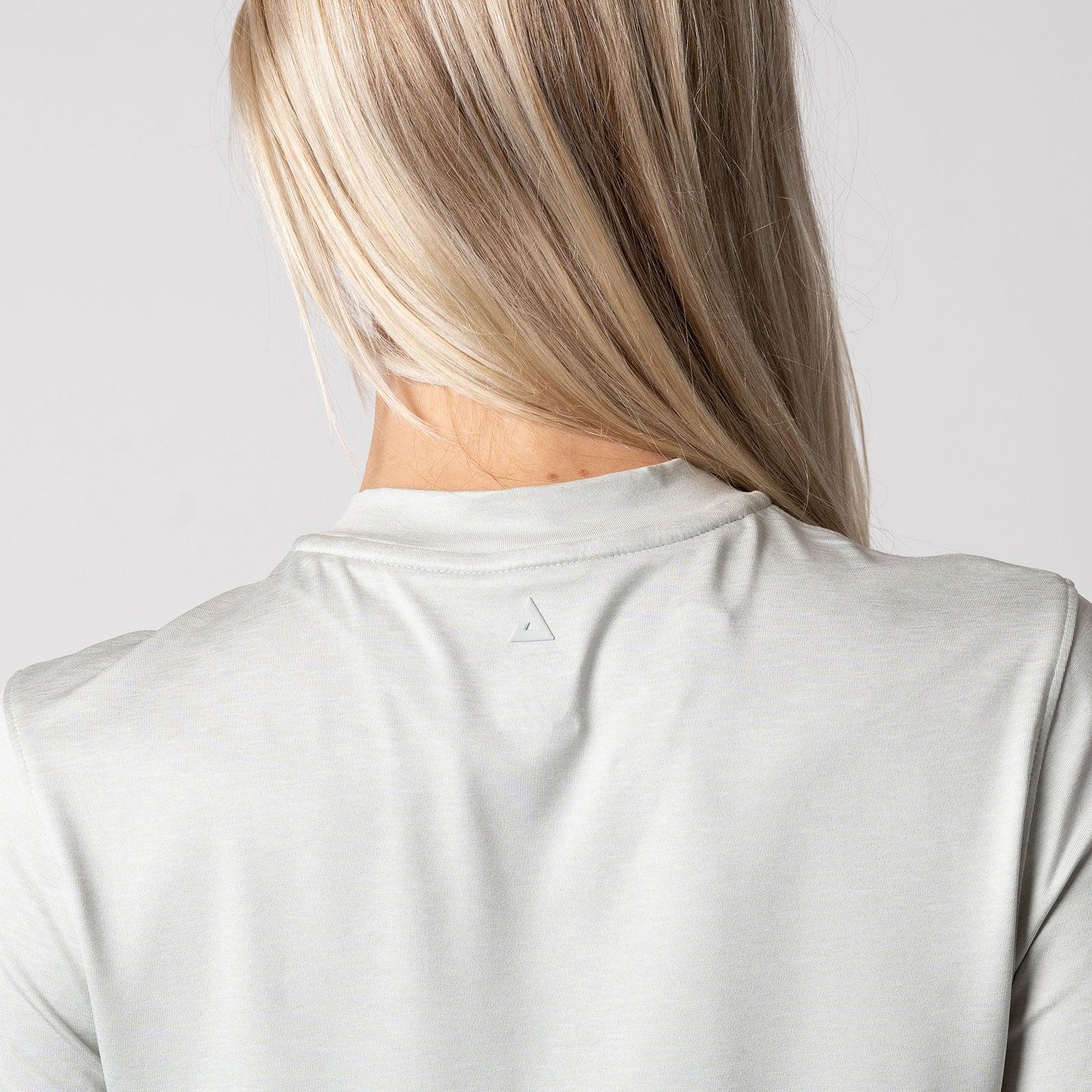 Frau mit langen blonden Haaren trägt ein hellgraues T-Shirt, Rückansicht, Fokus auf Nackenbereich mit kleinem, dreieckigem Logo.
