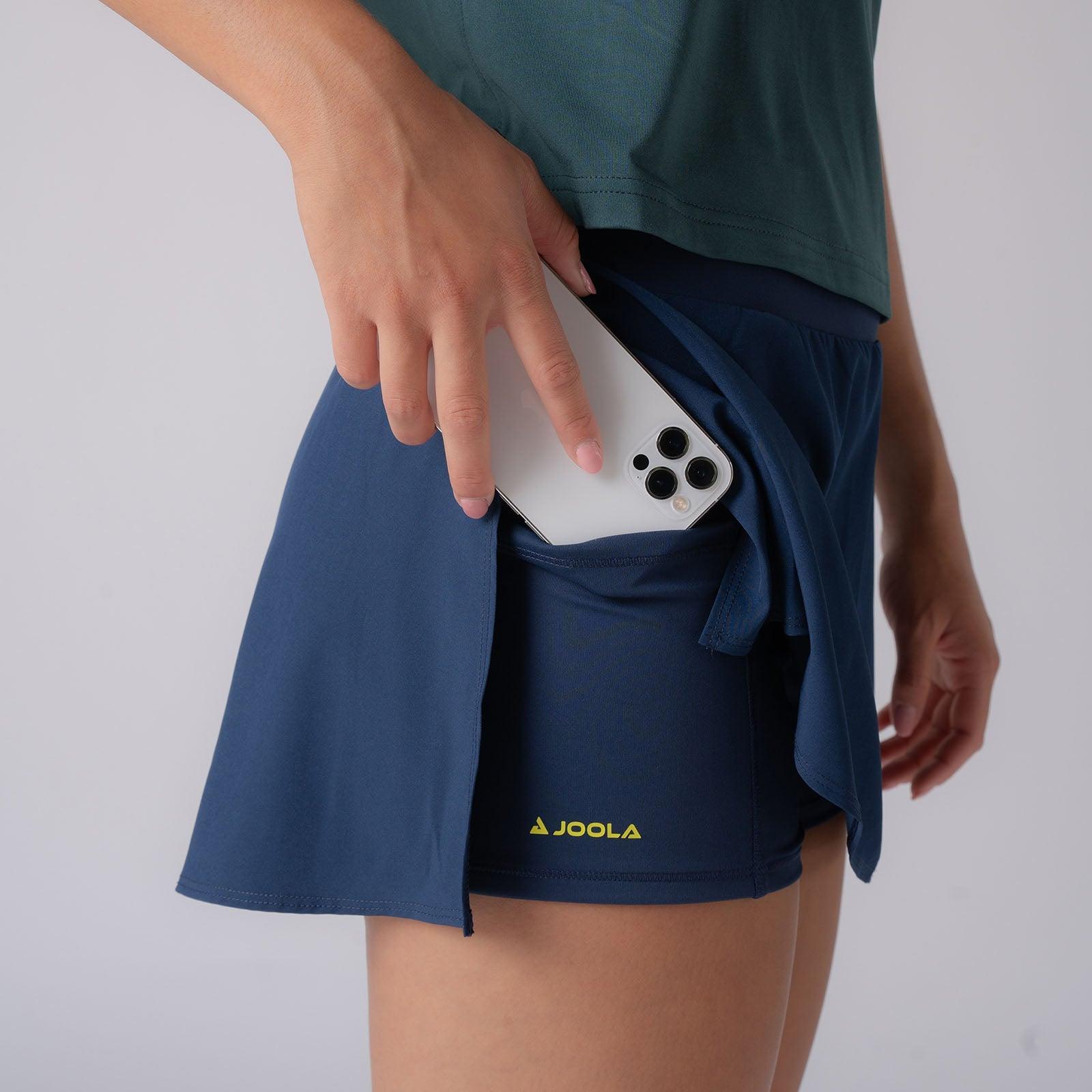 Frau steckt Smartphone in versteckte Tasche eines dunkelblauen Sportrocks mit Innenhose und gelbem JOOLA-Logo.