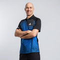 Mann trägt schwarzes und blau gestreiftes JOOLA Sport-Poloshirt, Arme verschränkt, lächelnd vor hellem Hintergrund.