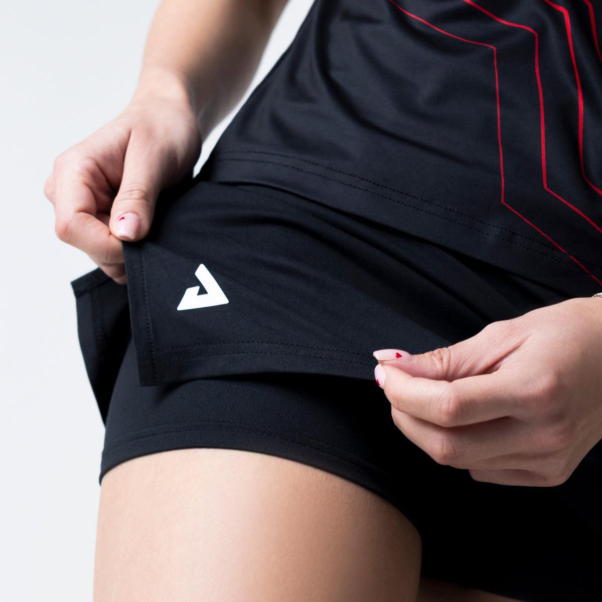 Frau hält schwarzen Sportrock mit integriertem kurzen Leggings, beide mit weißem Logo.