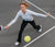 Frau in weißem Langarmshirt und schwarzer Hose spielt Pickleball und schlägt gelben Ball auf grauem Spielfeld.